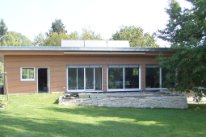 Einstöckiges Einfamilienhaus mit Holzfassade und Photovoltaikanlage auf dem Dach. Die grosse Fensterfront geht Richtung Garten.
